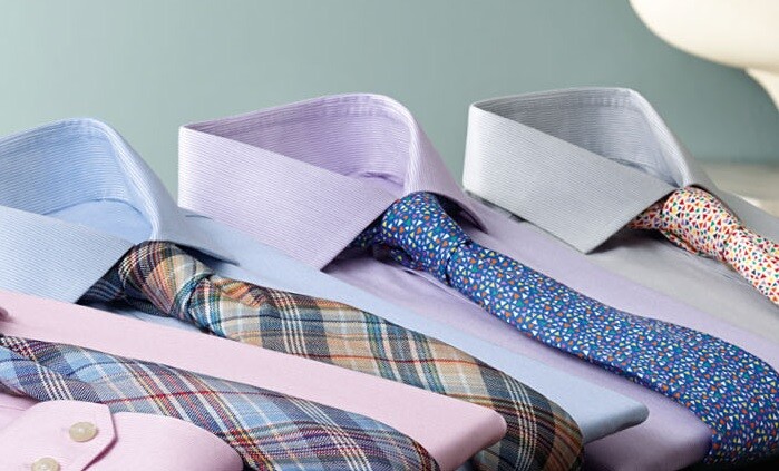 Four neatly folded dress shirts, each paired with a distinctive tie, are arranged in a row. The ties vary in patterns, including plaid, stripes, and multicolored dots.