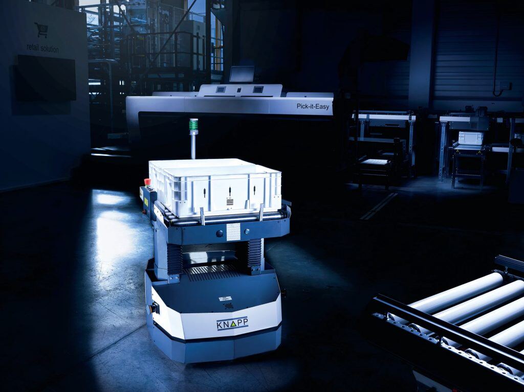 A robotic machine titled "KNAPP" transports a white container in a dimly lit industrial setting with various equipment and a "Pick-it-Easy" station in the background.