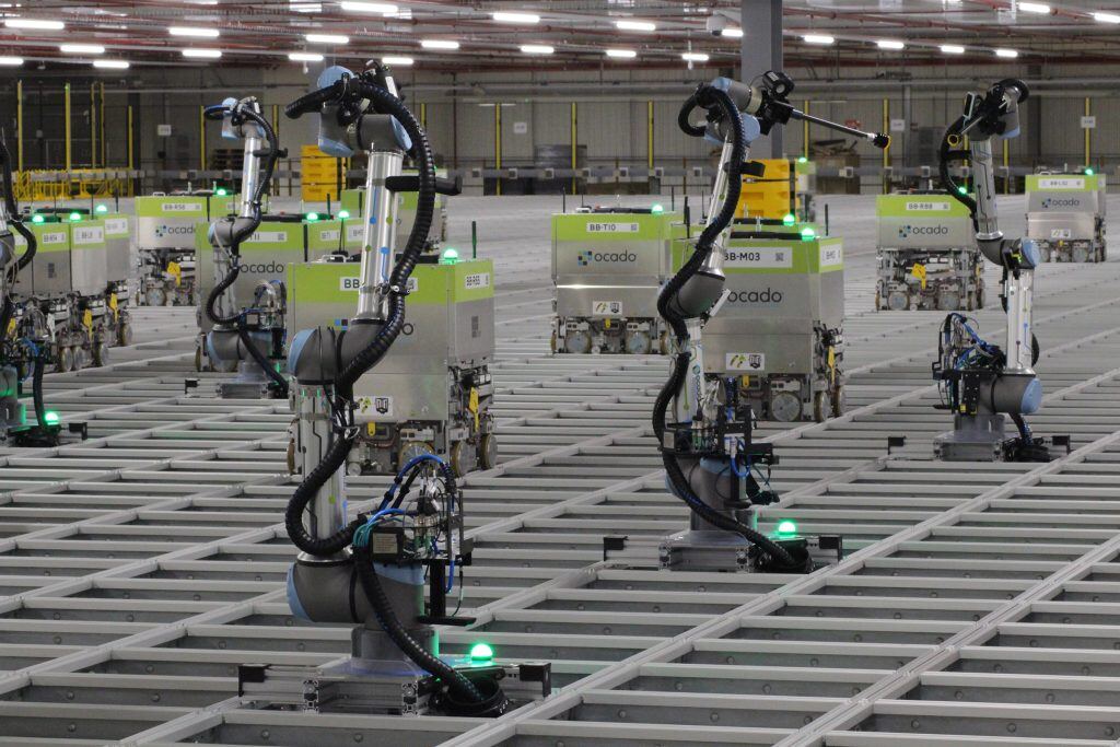 A warehouse features multiple robotic arms labeled "Ocado" positioned on a grid system, each equipped with various control units and green lights.