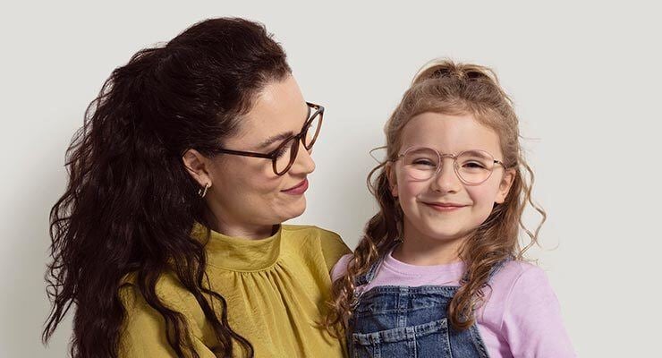 An adult with long, dark hair and glasses smiles at a child with curly hair and glasses, who is also smiling. The child is wearing a pink shirt and denim overalls.