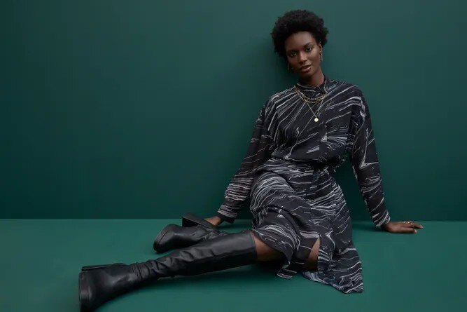 Person seated against a dark green background wearing a black and white patterned dress and black high boots.
